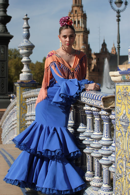 Descubre el Look Feria de Abril con el vestido de flamenca azul Jazmín, el mantón mandarina con flor buganvilla, ramillete Jaleo y pendientes dorados Carmona. Un conjunto vibrante y elegante para tu look flamenco.