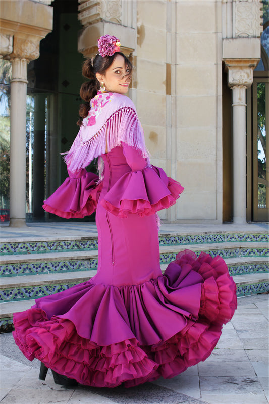 Deslumbra en la Feria de Abril con este vibrante Look Flamenco. El vestido de flamenca buganvilla se complementa con un mantón de seda lila con flor buganvilla y amarillo, ramillete Jaleo y pendientes dorados Nerja. Un conjunto que exuda pasión y elegancia.