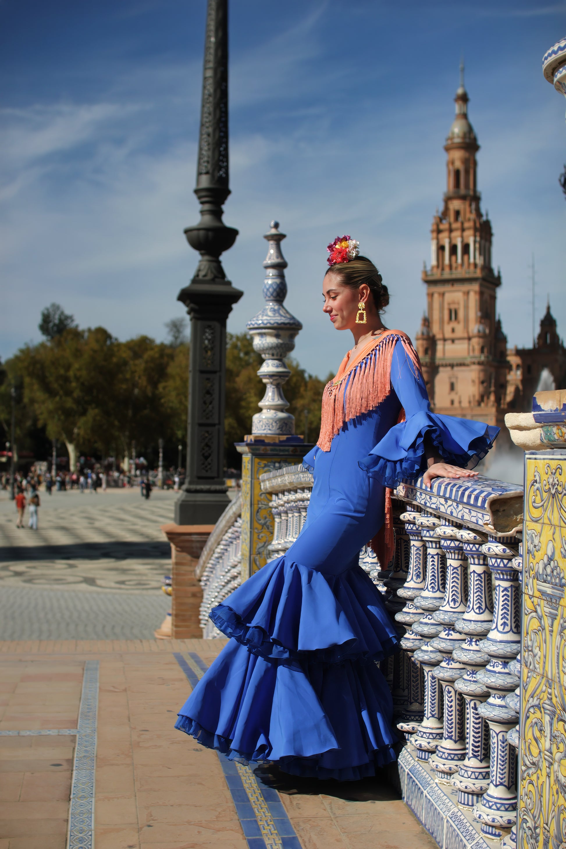 Destaca en la Feria de Abril con el vestido de flamenca azul Jazmin, manton mandarina con flor buganvilla, ramillete Jaleo y pendientes dorados Carmona. Un conjunto único y sofisticado.